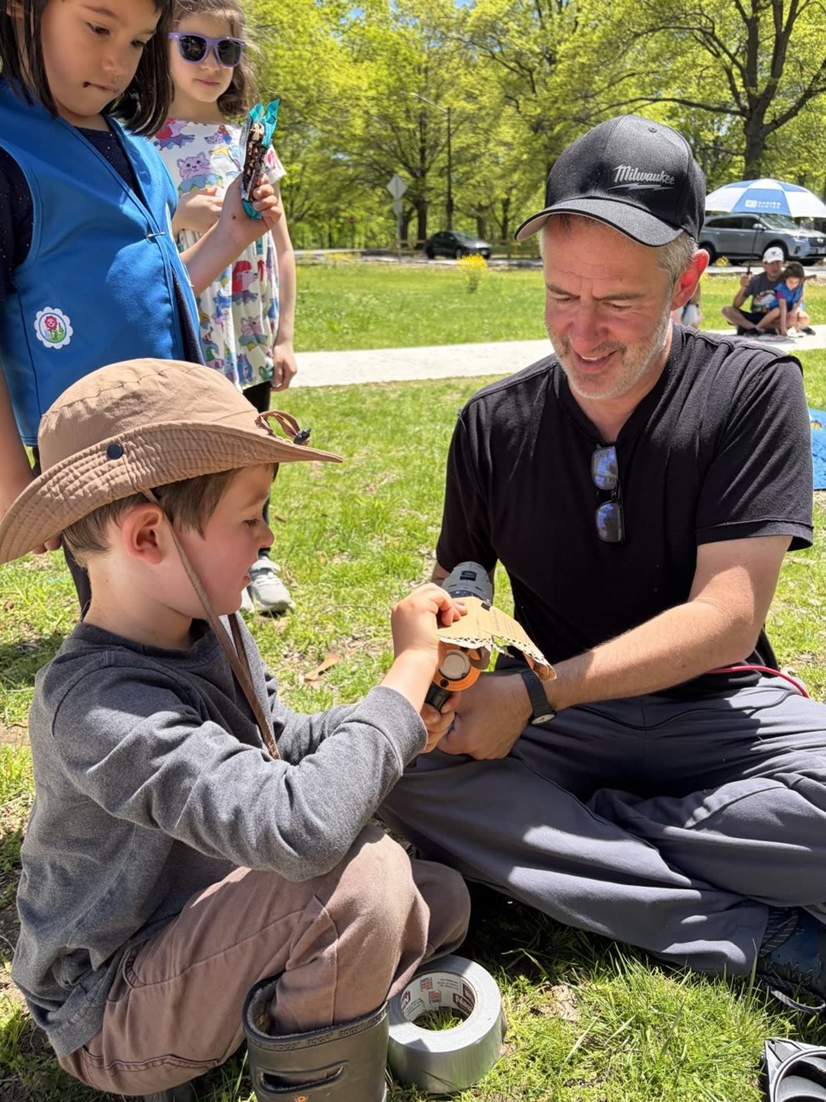 A young workshop participant in a brimmed hat sitting on the grass with an instructor, assembling a small electronics project together while two Girl Scouts look on