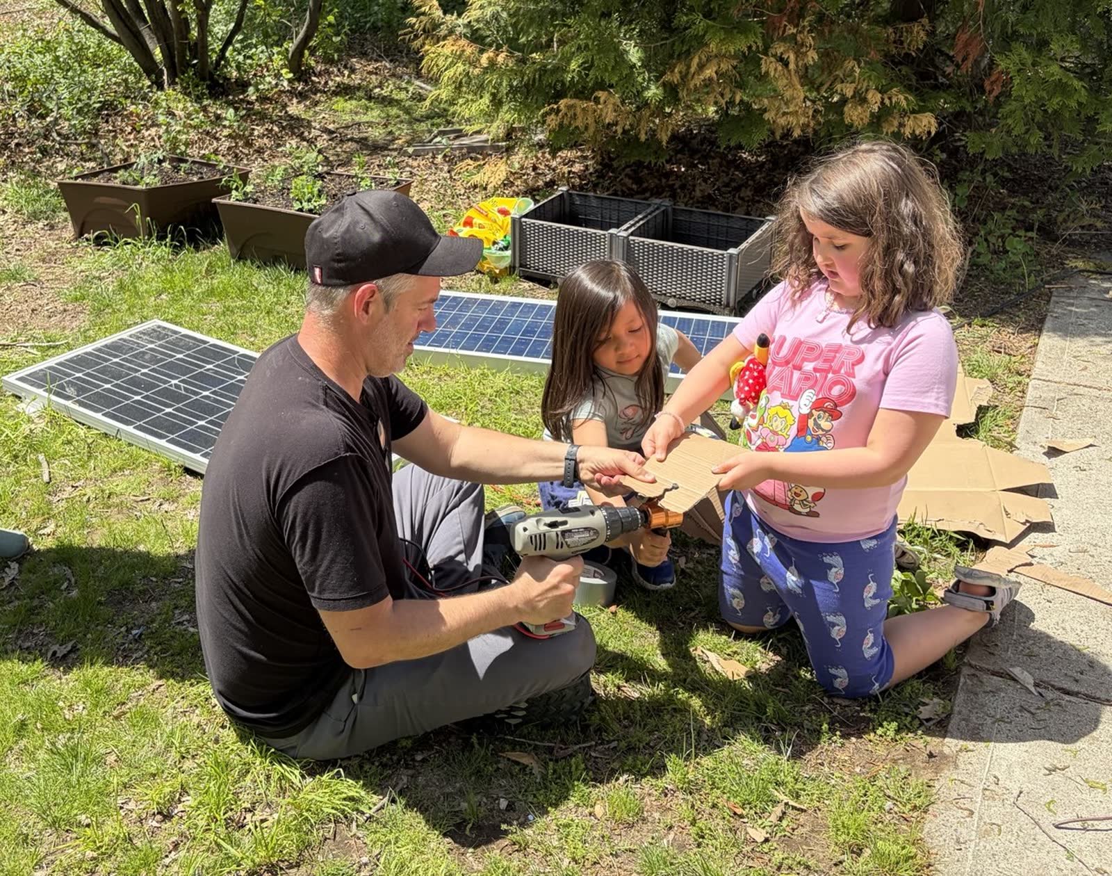 Two Girl Scouts and an instructor crouched on the grass cutting a piece of cardboard with a solar-powered nibbler; a small solar panel propped behind them
