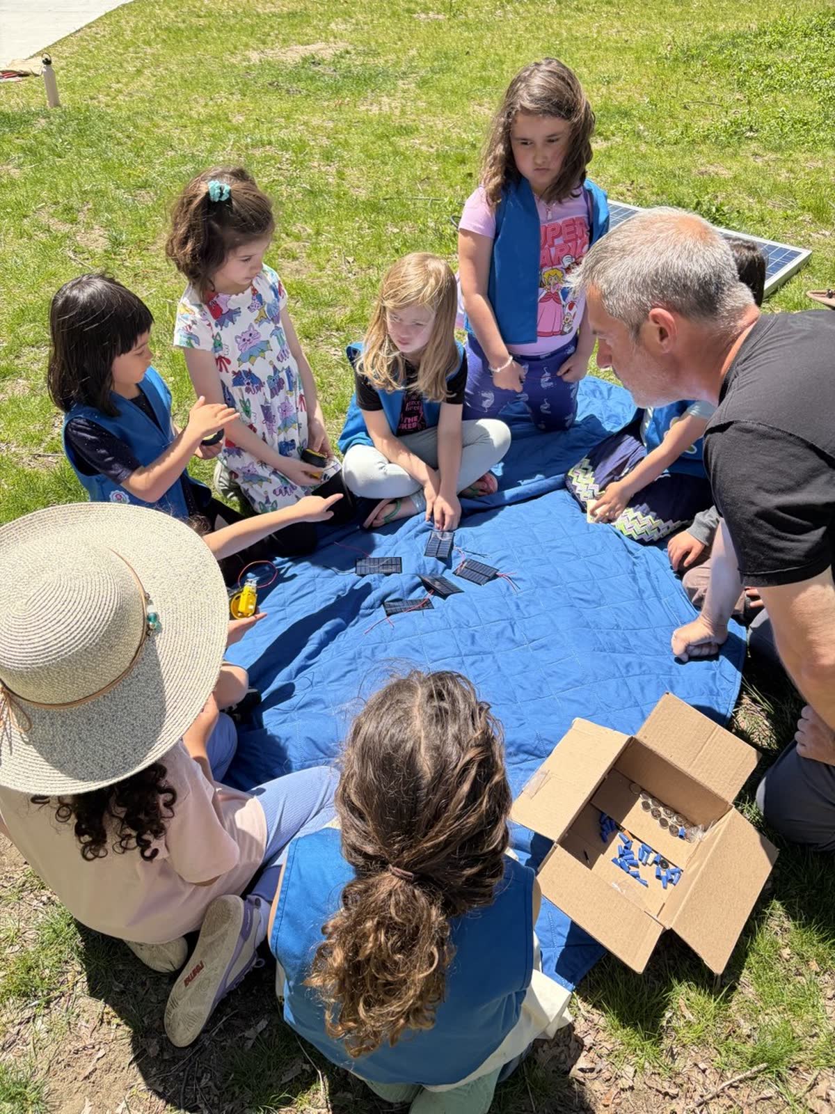 A Girl Scout troop gathered around a blue tarp examining small solar cells; a workshop instructor sits with them, a cardboard box of materials on the grass alongside